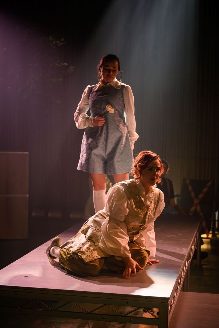 Two Opera Singers in white shirts with silk long tunics in a paisley and floral patterns. One singer is holding a white flower behind the other singer who is kneeling on the floor.